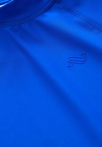 Modra športna majica iz gladkega, lahkega materiala. Ponaša se z nevsiljivim logotipom v nekoliko temnejši odtenek. Brezšivni vratni izrez in čisti robovi.