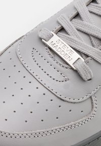 Graue Leder-Sneaker mit durchbrochenen Details, flachen grauen Schnürsenkeln und einem Metalletikett mit der Aufschrift "Steve Madden". Glatte Textur und Gummisohle.