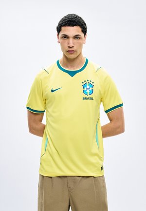 Νέος άνδρας στέκεται με τα χέρια πίσω από την πλάτη φορώντας κίτρινη φανέλα της Βραζιλίας με πράσινη λεπτομέρεια και έμβλημα.