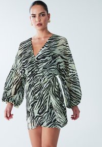 Mouwloze mini-jurk in doorschijnende mintgroene stof met zwart en gouden zebra print, voorzien van een diepe V-hals en een strikdetail bij de taille.