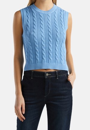 Frau trägt ärmelloses hellblaues Strickweste mit Zopfmuster und dunkelblaue Jeans, Hand in der Tasche, vor weißem Hintergrund.