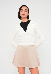 Λευκό πουλόβερ με πλέξη καλώδιο, V-λαιμόκοψη και μακριά μανίκια, σε συνδυασμό με μια μπεζ φούστα κοντή και φουσκωτή. Λείος υφασμάτινος και εφαρμοστός σχεδιασμός.