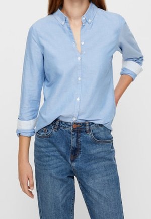 Femme portant une chemise bleu clair à boutons avec les manches retroussées et un jean en denim bleu, debout avec une main derrière le dos.