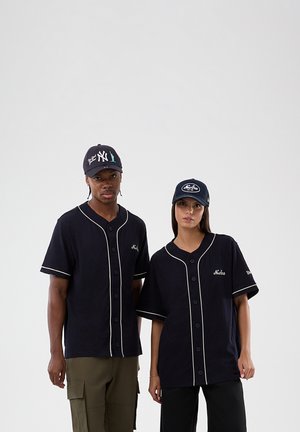Twee volwassenen dragen zwarte honkbalshirts met witte biezen en New Era-petten, staand tegen een effen lichte achtergrond.
