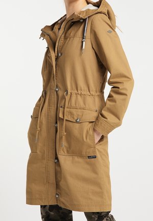 Mujer con una parka camel marrón con capucha hasta la rodilla, con cordones, cierres de botones y cremallera, y bolsillos delanteros con solapa, de pie con una mano en el bolsillo.