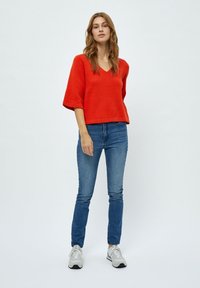 Maglione rosso a maniche corte con scollo a V, abbinato a jeans blu. L'outfit è completato da sneakers grigie. Design semplice e moderno.