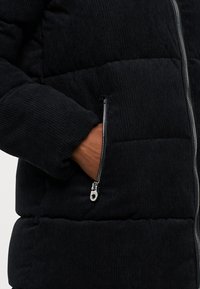 Juodas kordūrinis pūkų švarkas su užtrauktuku kišene, išryškinta raižyta tekstūra ir prigludęs kirpimas aplink apykaklę ir rankoves.
