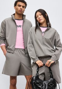 Δύο μοντέλα με ταιριαστά γκρι αθλητικά μπουφάν και σορτς, ροζ πουκάμισα από κάτω, το ένα κρατάει μια μαύρη δερμάτινη τσάντα, ουδέτερο φόντο.