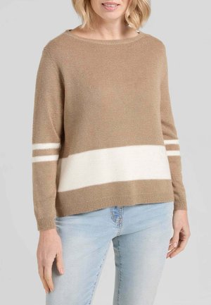 Donna che indossa un maglione beige con righe orizzontali bianche sulle maniche e sul torso, abbinato a jeans azzurri su uno sfondo semplice.