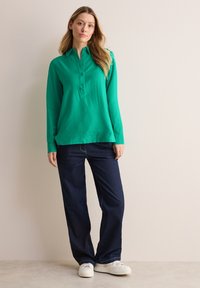 Zielona koszulka polo z długim rękawem, z zawadiackim kołnierzykiem, w zestawie z ciemnoniebieskimi spodniami dzwonami i białymi trampkami. Gładki materiał i luźny krój.