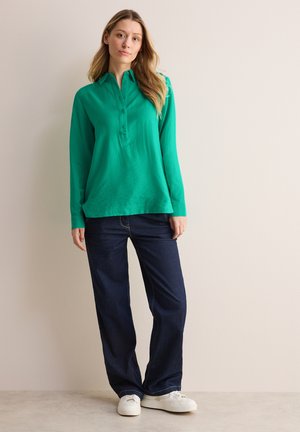 Groene lange mouwen polo met een knoopkraag, gecombineerd met donkere blauwe wijde jeans en witte sneakers. Gladde stof en een comfortabele pasvorm.