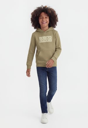 Enfant souriant aux cheveux bouclés marchant, portant un sweat à capuche vert olive Jack & Jones, un jean foncé et des baskets blanches sur fond blanc.