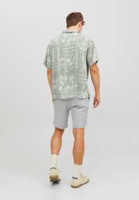 Camisa de manga corta verde claro con un patrón, pantalones cortos grises, zapatos beiges y calcetines blancos. La camisa presenta intrincados diseños blancos y un corte relajado.
