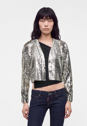 Jonge vrouw met lang zwart haar, die een zilveren paillettenjas, een zwart cropped topje en donkerblauwe spijkerbroek draagt, staand tegen een effen achtergrond.