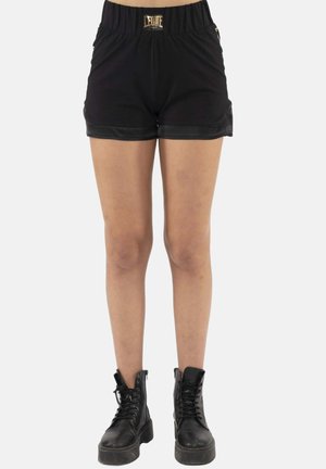Person trägt schwarze elastische Shorts mit goldenem Logo-Patch und schwarze Schnür-Stiefeletten, steht vor einfachem Hintergrund.