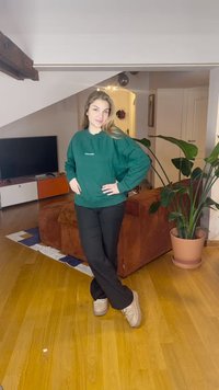 Zielona bluza z białym napisem, czarne spodnie i jasne buty. Pokój z drewnianą podłogą i doniczką z rośliną w tle.