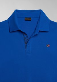Polo bleu avec un col, deux boutons partiellement déboutonnés, un petit logo de drapeau rouge et blanc sur la poitrine, et une étiquette de marque à l'intérieur du col.