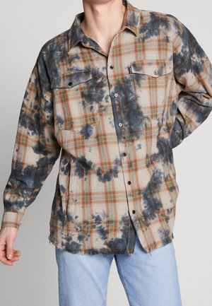 Mand iført en beige og orange ternet skjorte med sorte batikpletter og lyseblå jeans, der står foran en ensfarvet baggrund.