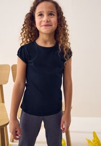 Schwarzes Kurzarm-T-Shirt mit Rundhalsausschnitt und gerafftem Schulterdetail, kombiniert mit grauen Leggings. Weicher Stoff, lässiger Schnitt, minimales Design.