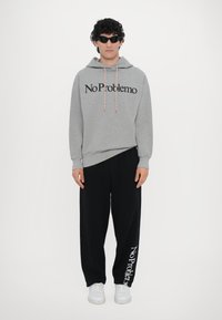 Grauer Baumwoll-Hoodie mit dem Aufdruck "No Problemo" in Schwarz, kombiniert mit schwarzen Jogginghosen mit Logo-Print. Das Modell trägt Sonnenbrillen und weiße Sneakers.