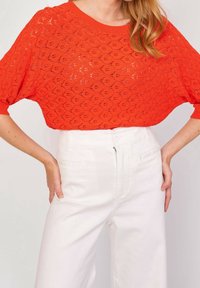 Nainen, jolla on kirkkaan oranssi virkatulla lyhythihainen paita ja vyötäröltä korkeat valkoiset housut, kädet lepäävät lanteilla.