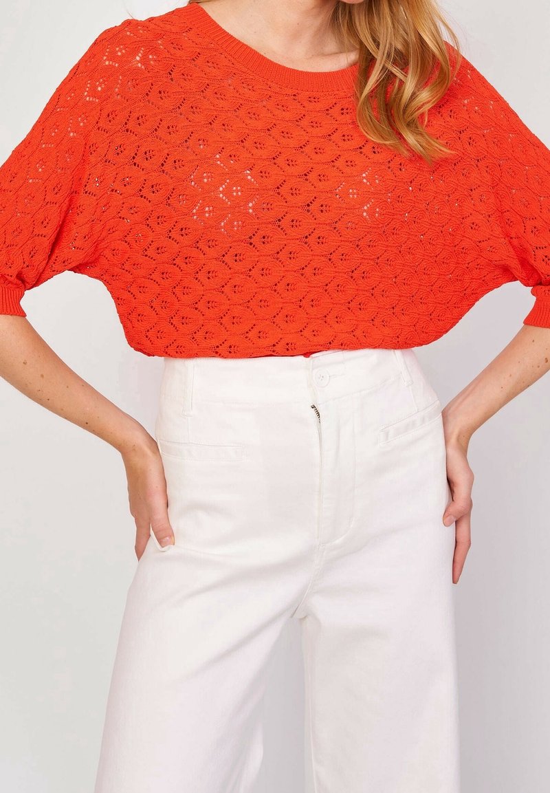 Nainen, jolla on kirkkaan oranssi virkatulla lyhythihainen paita ja vyötäröltä korkeat valkoiset housut, kädet lepäävät lanteilla.