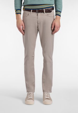 Mand iført beige smalle bukser, lyse beige sneakers og en grøn sweater med stribede manchetter og kant, stående mod en hvid baggrund.