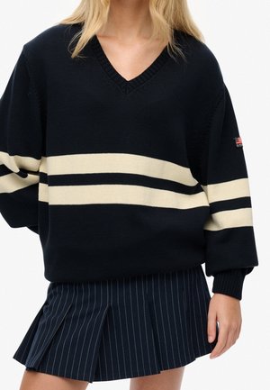 Person iført en sort V-hals sweater med to hvide vandrette striber og et mørkt plisseret skørt med hvide lodrette nålestriber.