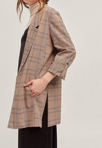 Hnědý kostkovaný blejzr s dlouhými rukávy, který má volný střih, náprsní kapsy a klopové límce, vyrobený z texturované látky.