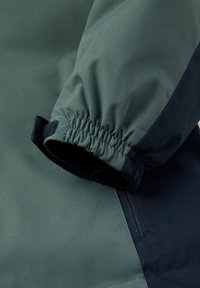 Groene jasvoering met elastische manchetten, voorzien van een donkerblauwe accenten. Glad, lichtgewicht stof met een stevige klittenbandsluiting.