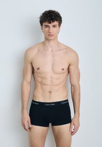 Boxer briefs neri con il logo Calvin Klein sulla cintura; il modello ha un fisico tonico e capelli corti e ricci, in posa su uno sfondo neutro.