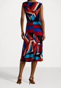 Mujer que lleva un vestido sin mangas, de corte medio, con audaces patrones geométricos abstractos en rojo, azul, negro y crema, combinado con sandalias de tacón alto marrones.