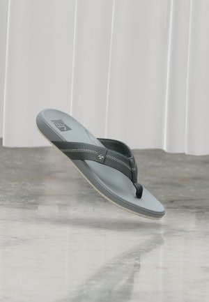 Sandale tongs grise et noire avec semelle intérieure texturée, couture en zigzag sur les brides, et logo sur la semelle, suspendue au-dessus d’un sol poli.