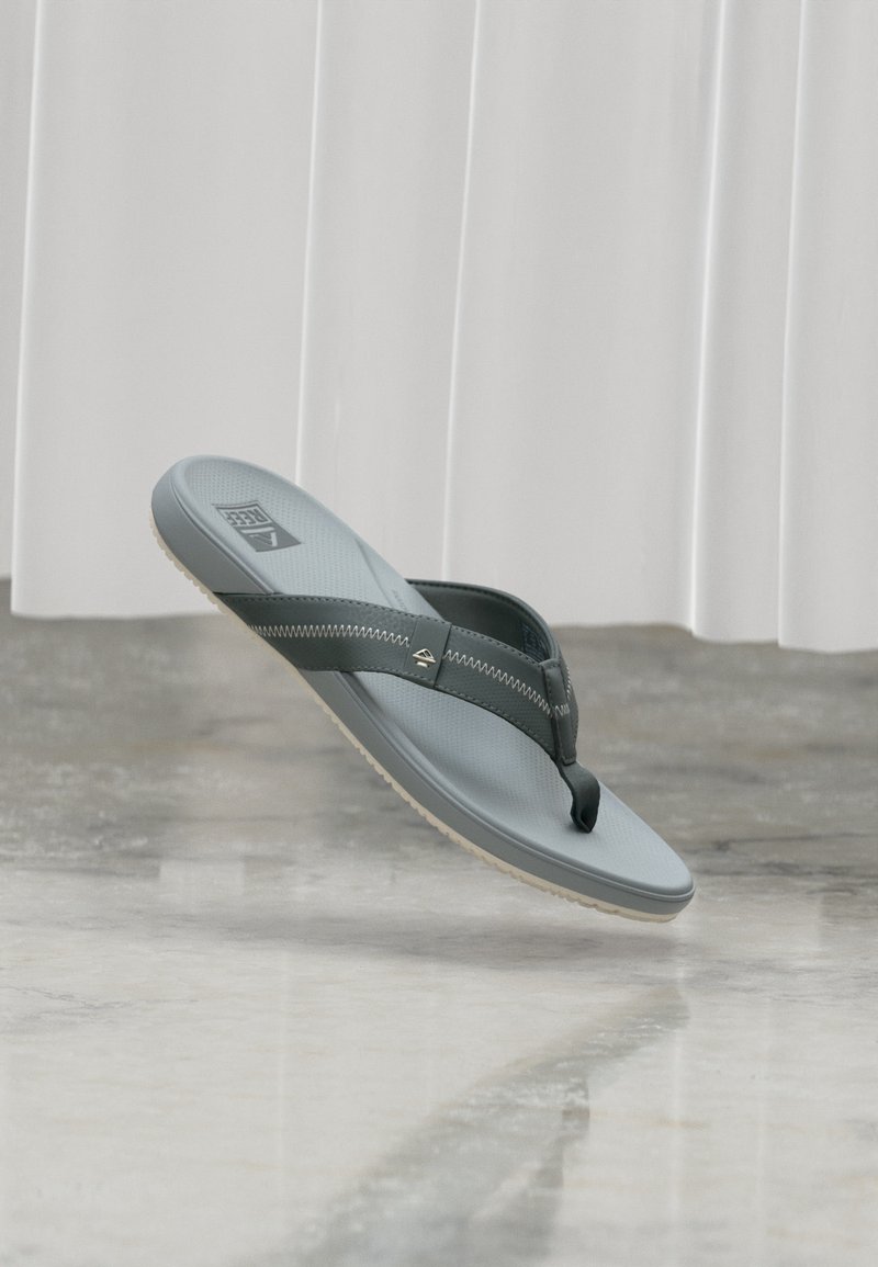 Γκρι και μαύρο σανδάλι τύπου φλιπ-φλοπ με υφή στο πέλμα, ραφή ζιγκ-ζαγκ στα λουριά και λογότυπο στην εσωτερική σόλα, κρεμασμένο πάνω από γυαλισμένο δάπεδο.