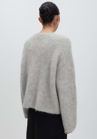 Vrouw met haar in een knot die zilveren hoepeloorbellen en een lichtgrijze fuzzy trui draagt, gezien van achteren op een effen achtergrond.