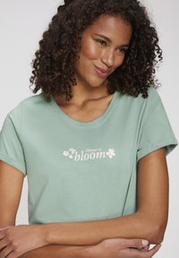 Mentolovo zelené tričko, krátke rukávy, lodičkový výstrih. Obsahuje text "vyber si kvitnúť" s kvetinovými akcentmi v bielej farbe. Mäkká, pohodlná tkanina.