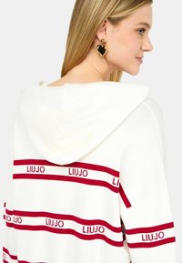 Felpa bianca con strisce rosse e logo "LIUJO". Tessuto morbido, design casual e cappuccio. Orecchini a forma di cuore dorati indossati.