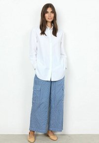 Camicia bianca con colletto, abbinata a pantaloni larghi a righe blu con tasche cargo. Ballerine beige completano l'outfit.