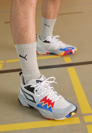 Zapatos deportivos blancos con acentos multicolores (rojo, azul, amarillo) y detalles en negro. Material texturizado, cordones gruesos y blancos, usados con calcetines grises.