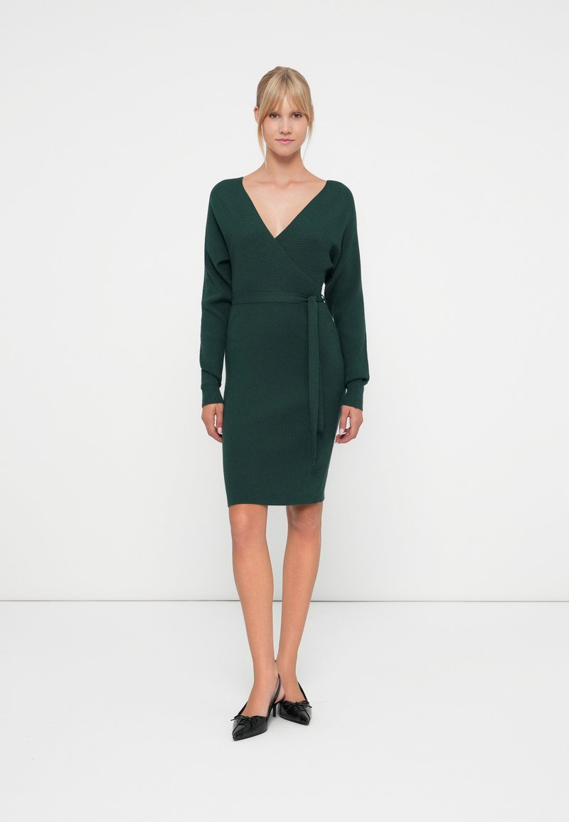 Robe en tricot verte avec un décolleté en V profond, des manches longues, un design enroulé et une ceinture à nouer à la taille. Chaussures noires à talons pointus.