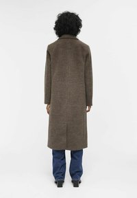 Lange bruine wollen jas met een kraag, recht gesneden ontwerp en zijspleten, gecombineerd met blauwe spijkerbroek en zwarte hakken. Effen achtergrond.