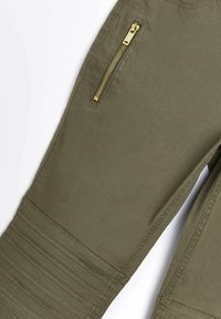 Olivgröna byxor med texturerad front, med en guldzippered ficka och vertikala sömmar på knäna.