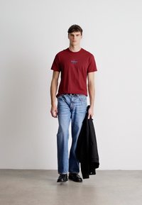 Burgundi t-särk sinise graafikaga, heledad sinised teksad, must jakk paremas käes ja mustad kingad. Ühtlane taust.