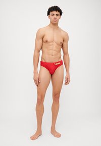 Slipuri de baie roșii dintr-un material elastic, cu un accent logo auriu pe talie. Textură netedă, design ajustat și acoperire minimă.
