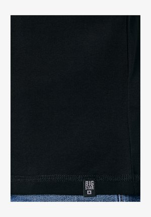 Fekete vászon látható varrásokkal és egy kis fekete címkével, amelyen fehér betűkkel a "BIG STAR" felirat olvasható, a kék farmer anyag aljánál varrva.