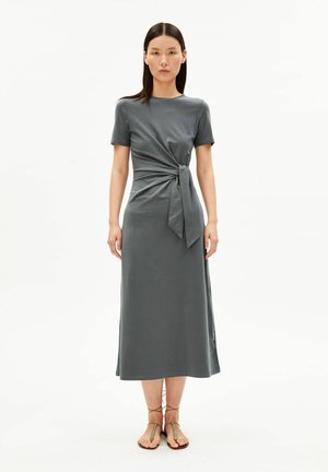 Woman with straight dark hair wearing a gray midi dress with side knot detail and lace-up flat sandals, standing against a white background.