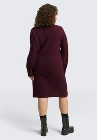 Burgunderfarbene Strickkleid mit langen Ärmeln, geradem Schnitt und minimalen Nahtdetails, kombiniert mit schwarzen Knöchelstiefeln.