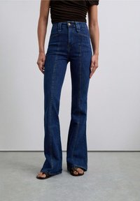 Jeans a vita alta a zampa d'elefante in denim blu scuro, caratterizzati da cuciture evidenti e una chiusura a bottone, abbinati a sandali scuri che mostrano le dita dei piedi.