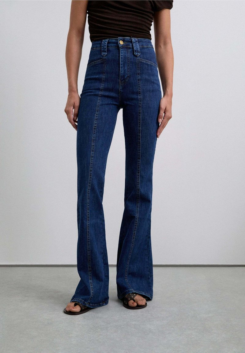 Jeans a vita alta a zampa d'elefante in denim blu scuro, caratterizzati da cuciture evidenti e una chiusura a bottone, abbinati a sandali scuri che mostrano le dita dei piedi.