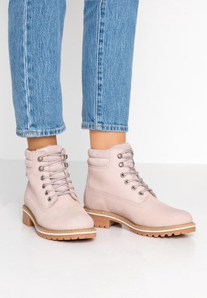 Jasnoróżowe sznurowane botki za kostkę z wyściełanym kołnierzem i brązowymi, grubymi podeszwami noszone z niebieskimi dżinsami.